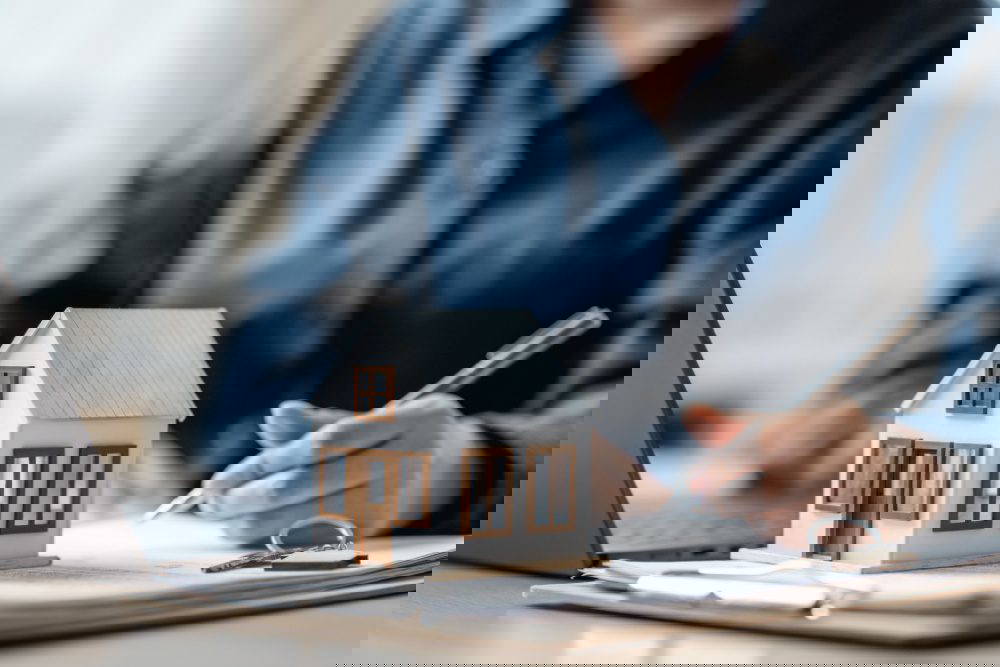 A person signing a document with a model house on the table.